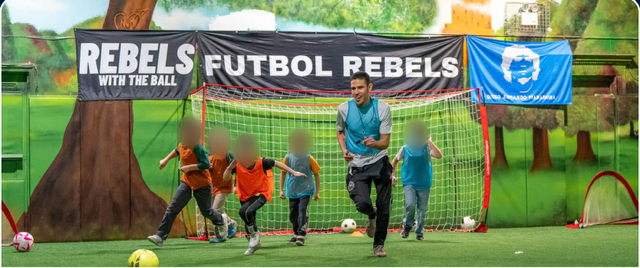 A Futbol Rebels coach running with young players on an indoor turf field